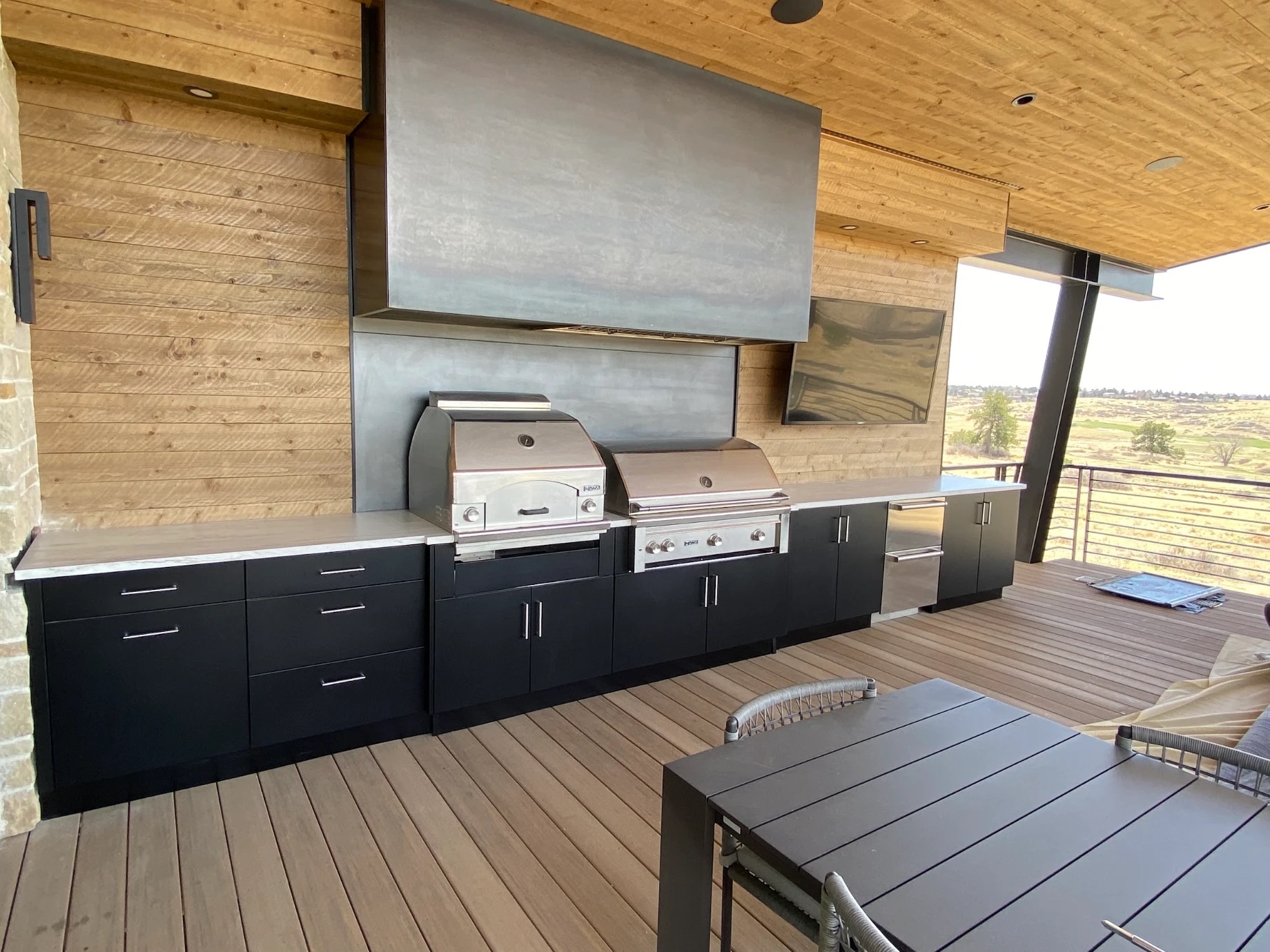 Black outdoor cabinets with a pizza oven and grill under a steel hood on a covered deck