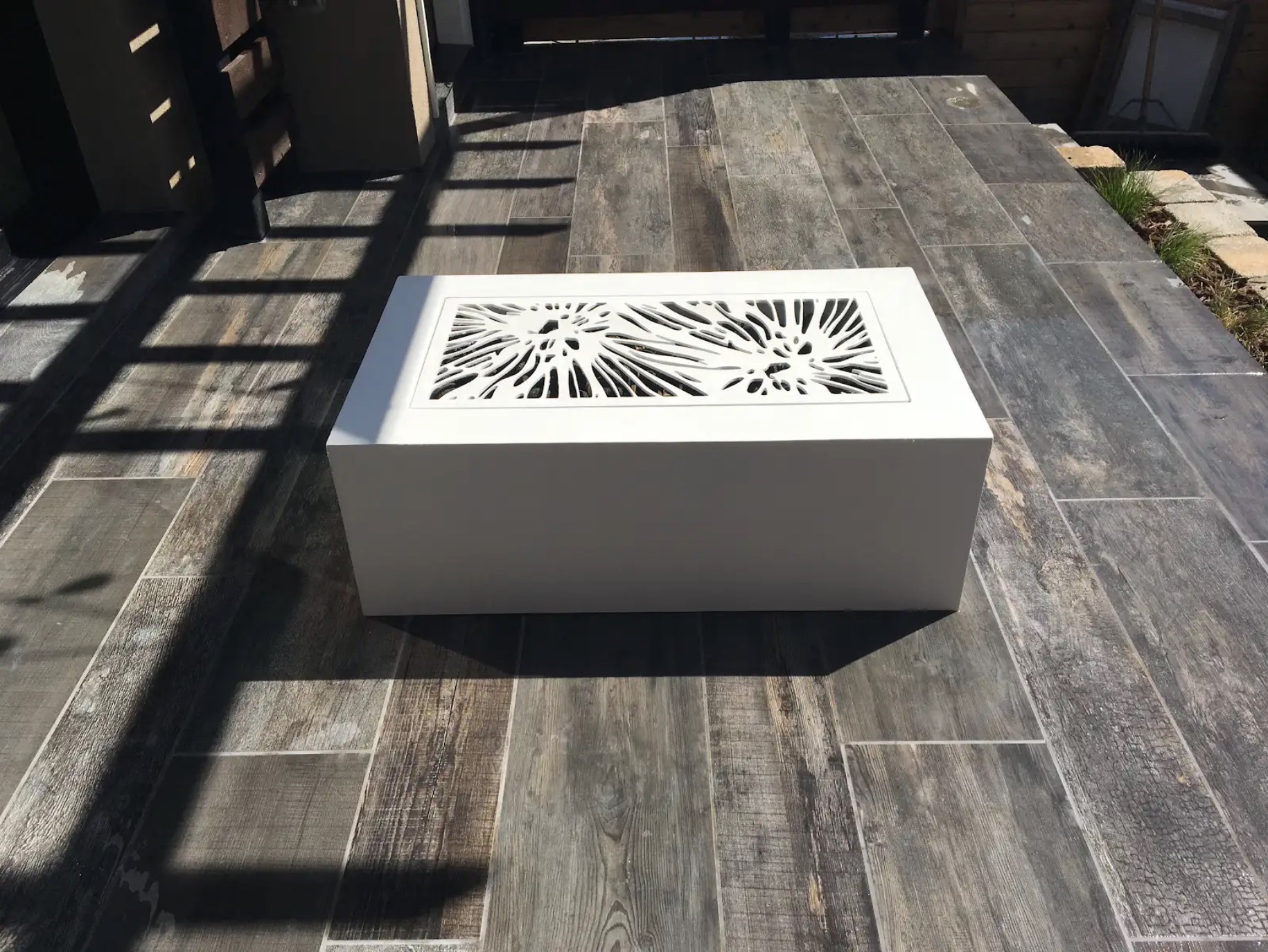White powder-coated steel firepit with a decorative laser-cut cover on a wood-look tile deck