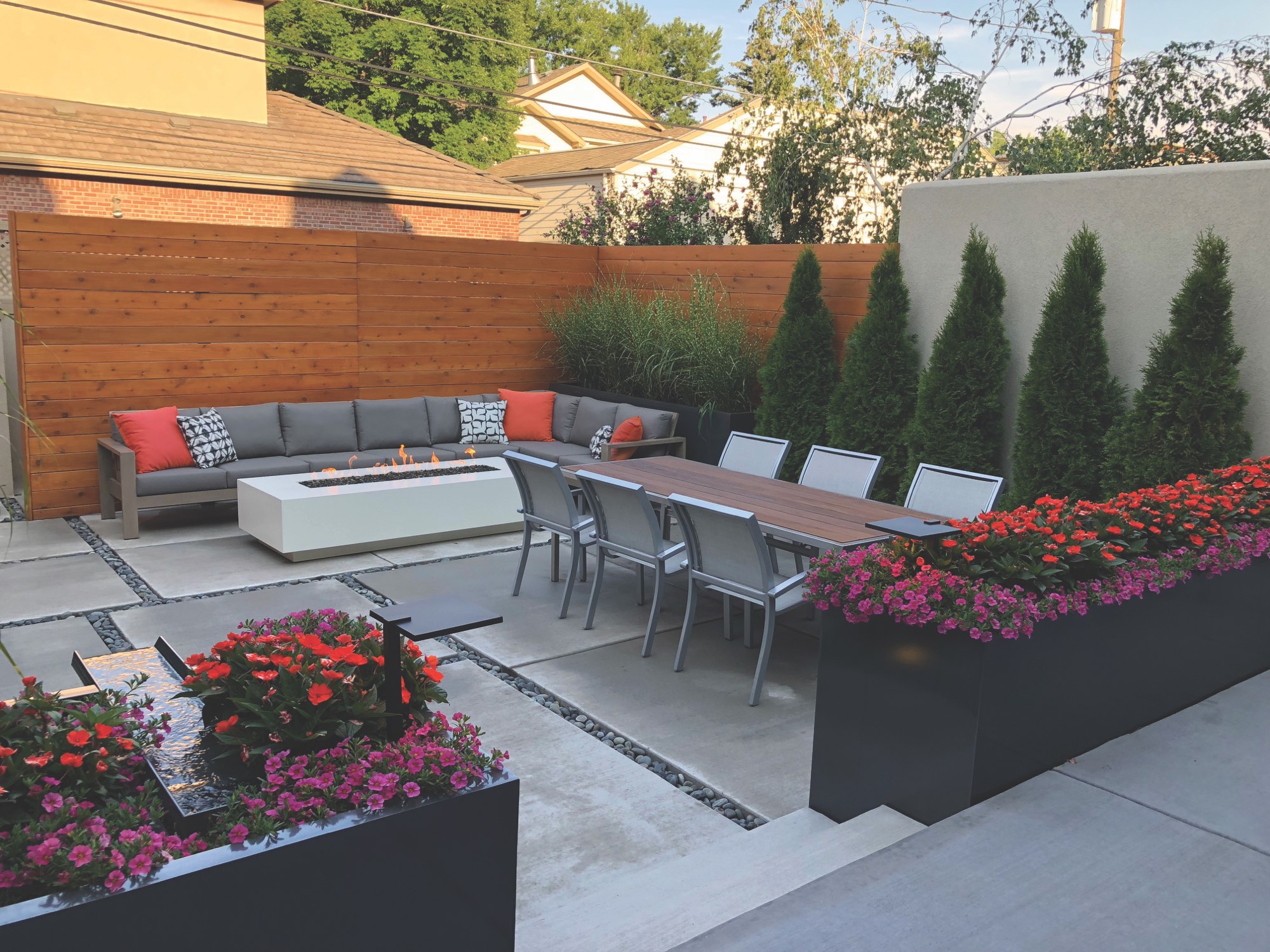 Long white steel firepit in a modern courtyard with a sectional sofa, dining table and flower planters
