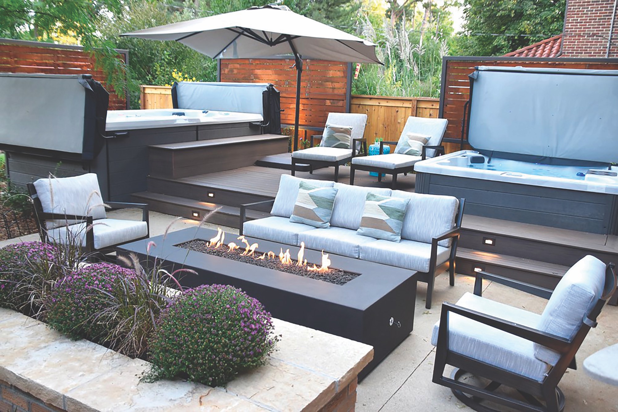 Long dark powder-coated steel linear firepit with a row of flames on a terraced patio beside lounge seating and hot tubs