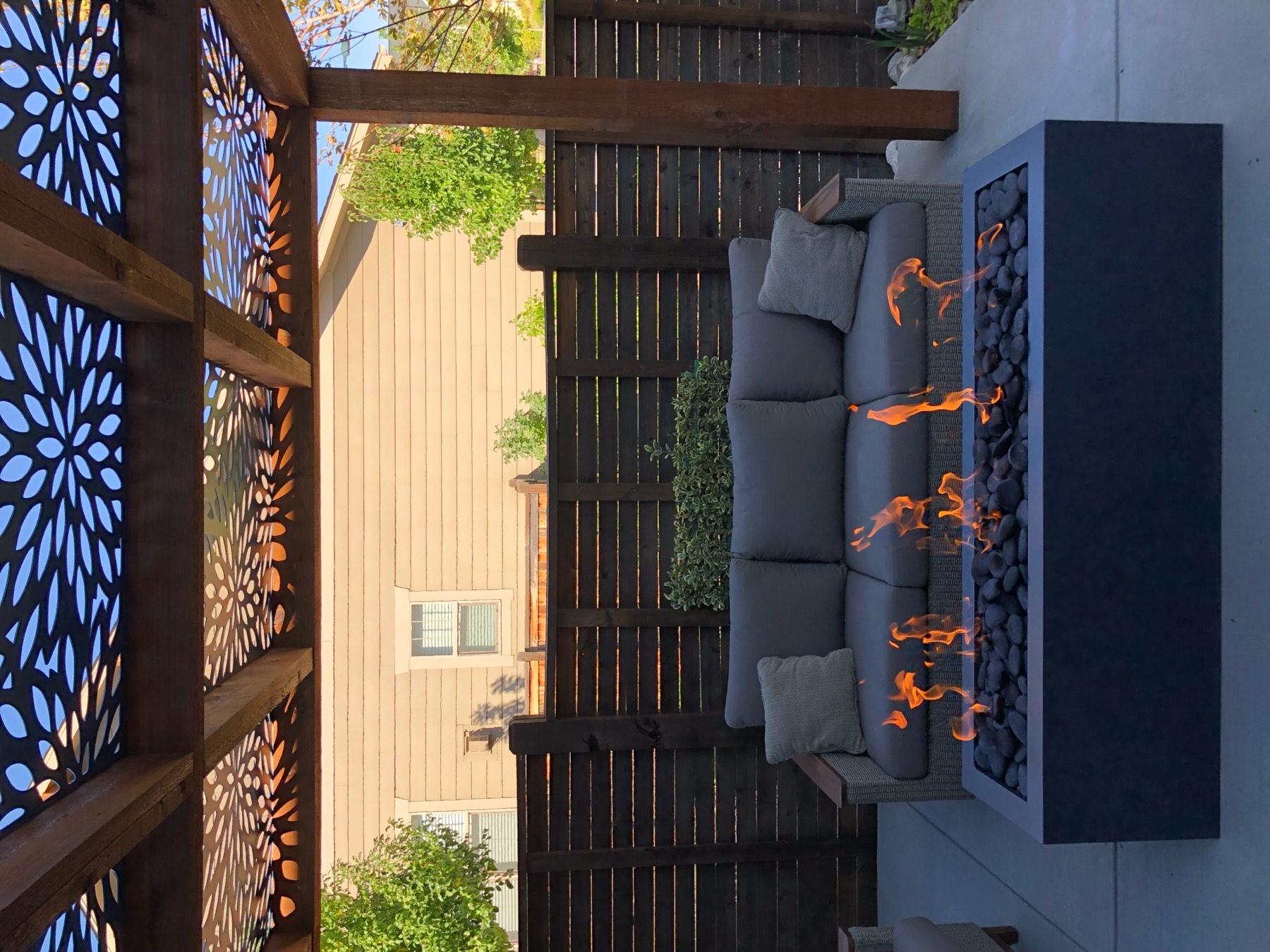 Dark rectangular steel firepit with flames under a pergola with a decorative cut-metal canopy