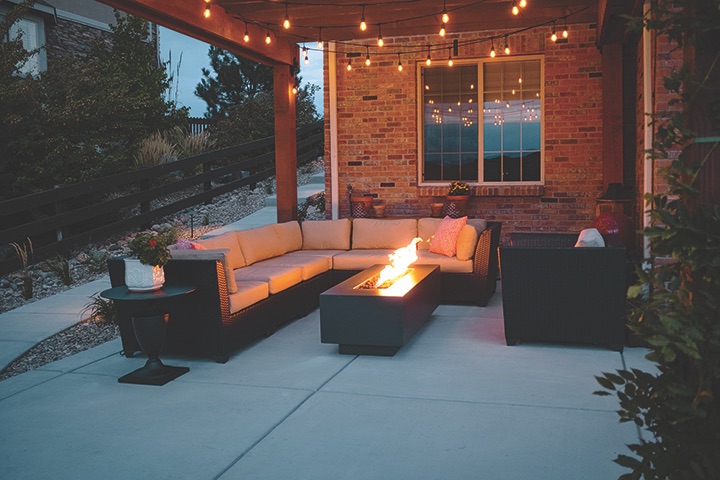 Dark linear steel firepit glowing at dusk on a covered patio with a sectional sofa and string lights