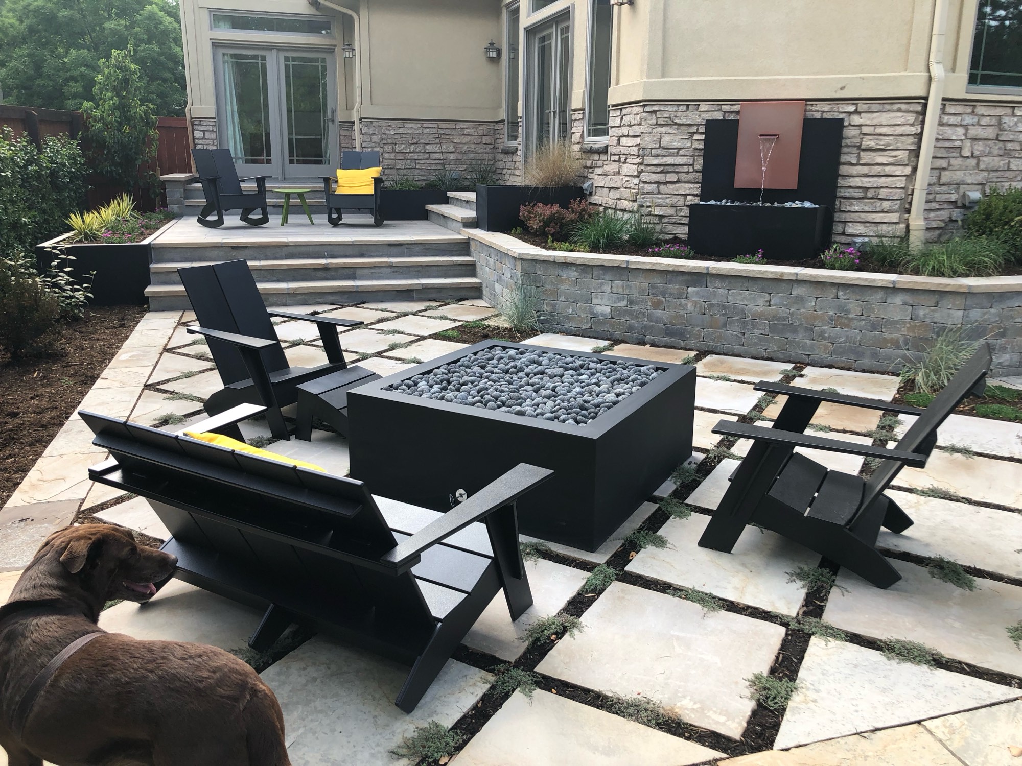 Dark square steel firepit filled with fire glass on a stone patio with lounge chairs