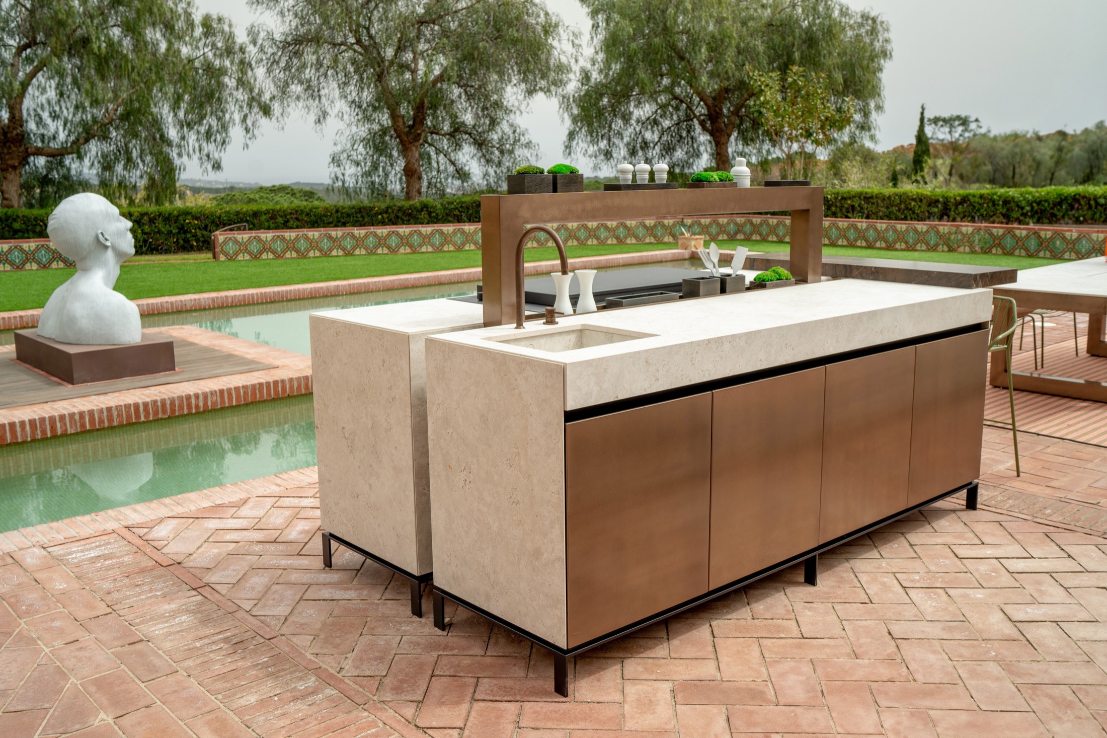 Novara island with a pale stone top and liquid metal bronze fronts beside a garden pool