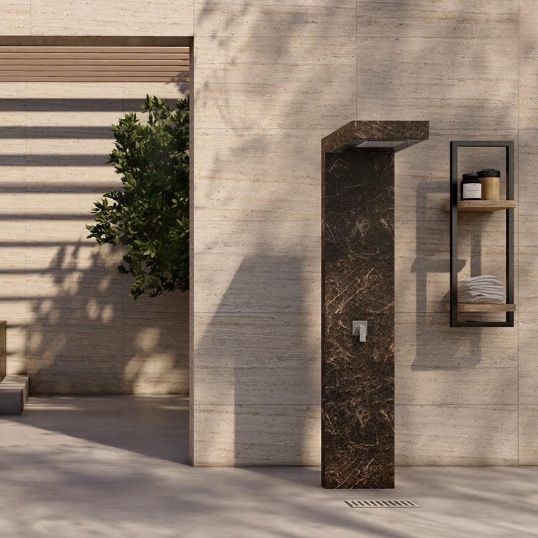 Korta column shower clad in dark marble against a travertine wall, beside a wall shelf with towels
