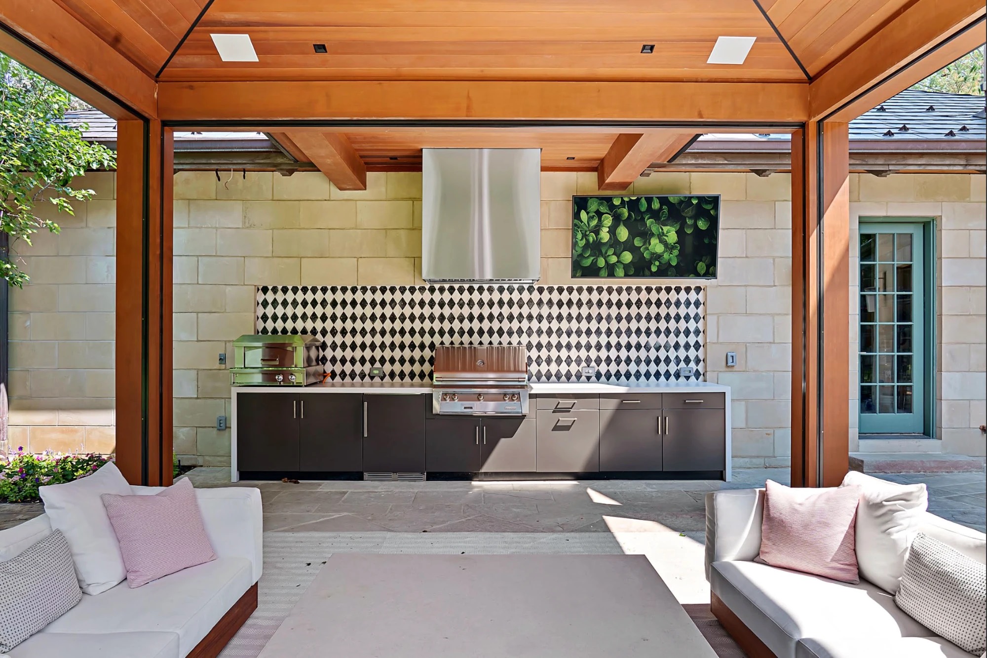 Dark outdoor kitchen with a waterfall countertop, pizza oven, grill and stainless hood under a cedar pavilion with a diamond-pattern backsplash
