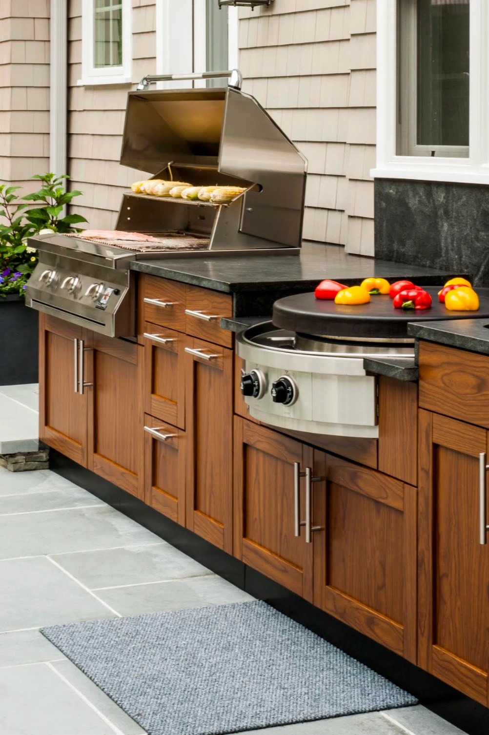Wood-grain outdoor cabinets with a stainless grill, a round griddle and a soapstone-look countertop beside a shingled house